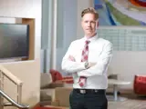 A business man wearing a white dress shirt, red plaid tie, and black trousers stands confidently with his arms crossed